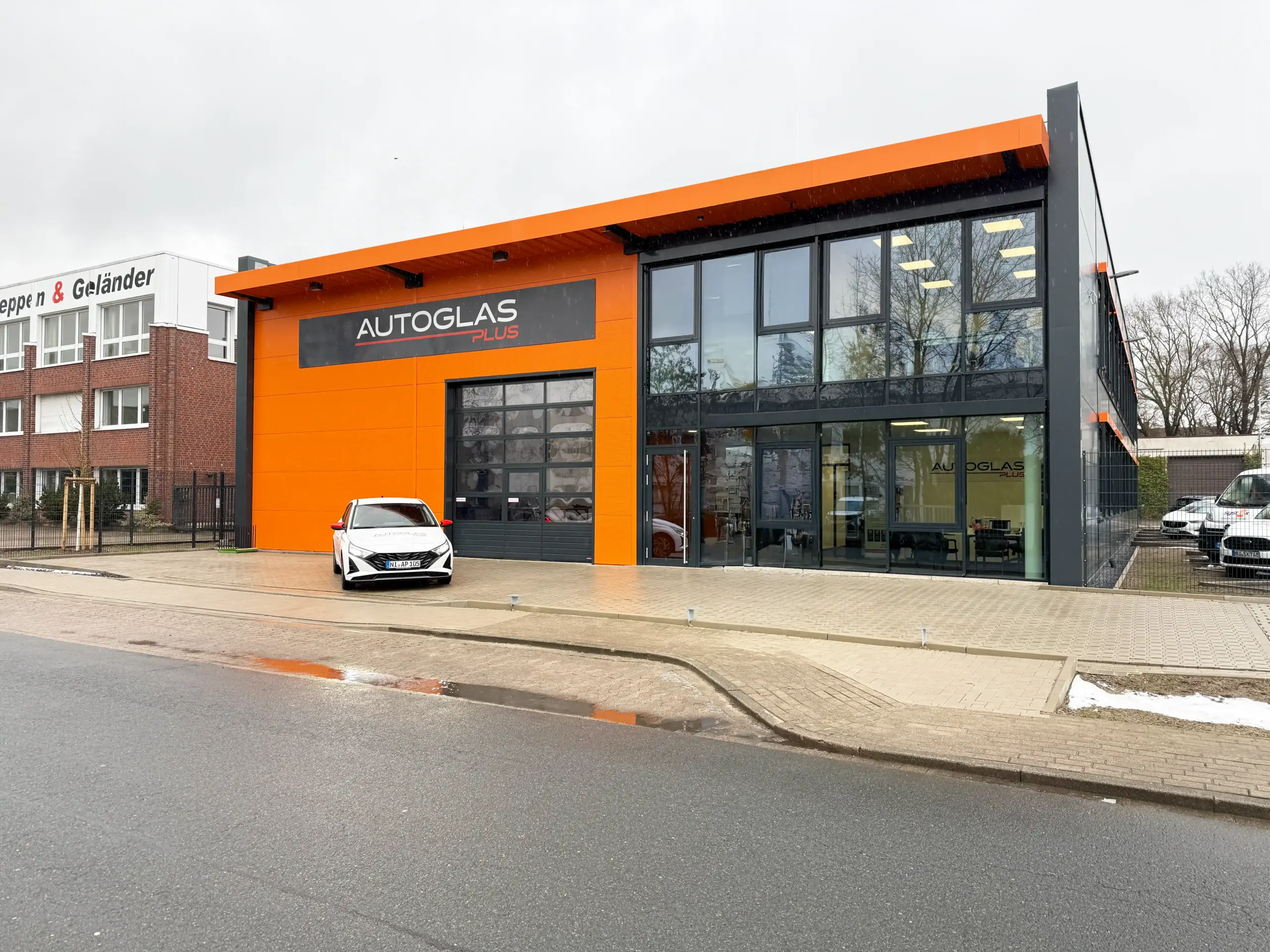 Ein modernes orange-schwarzes Gebäude mit der Aufschrift "AUTOGLAS PLUS" mit großen Fenstern, einem davor geparkten weißen Auto und einem wolkenverhangenen Himmel über uns.