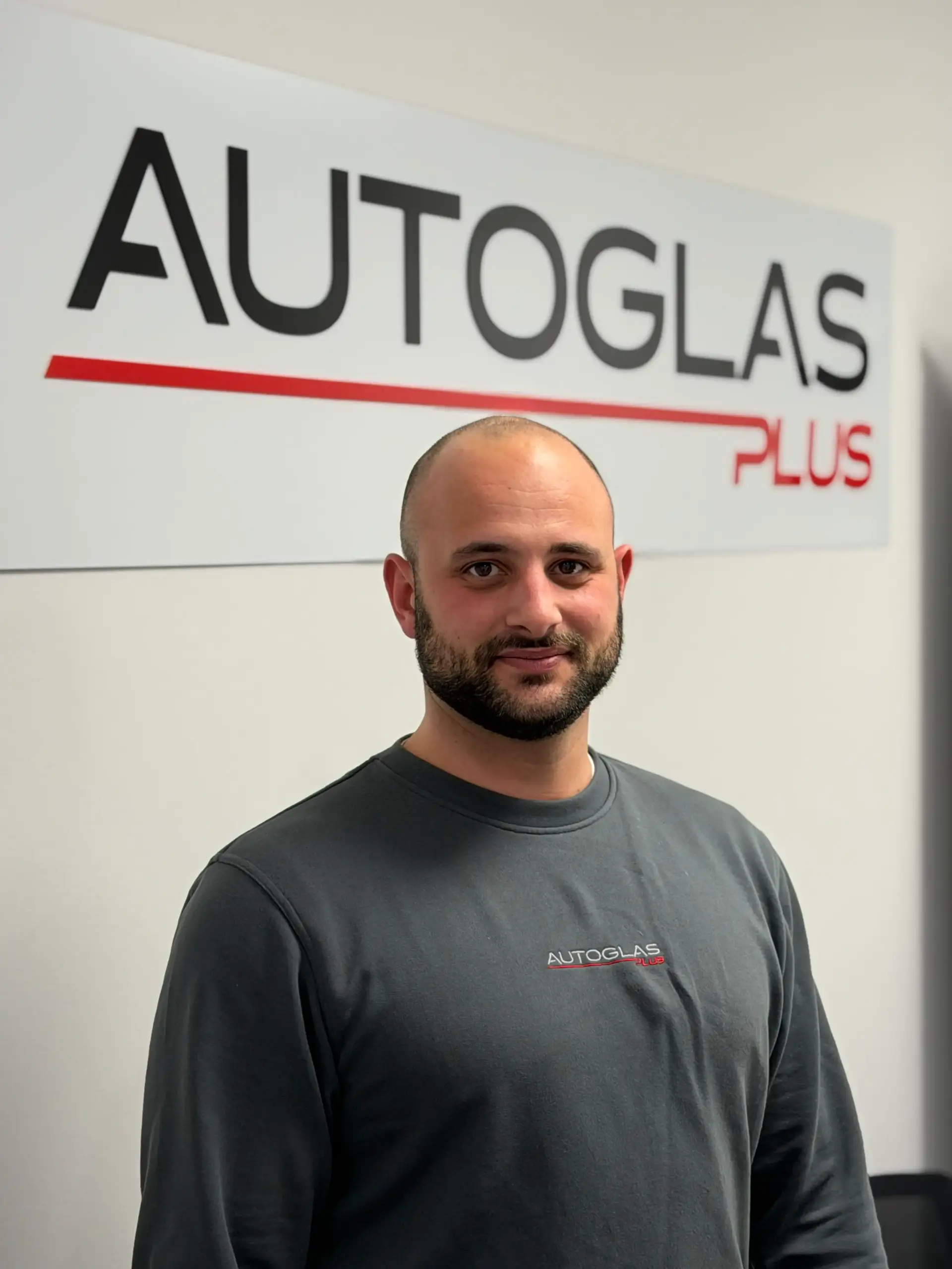 Autoglasplus_Hannover_Viktor_Bittel Ein Mann mit Bart steht vor einem "AUTOGLAS PLUS"-Schild und trägt ein graues Hemd mit demselben Logo.
