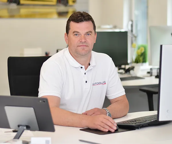 Steinschlagreparatur_Scheibentausch_Scheibentoenung_Bremen_autoglas-plus_Vitalij-Blank Ein Mann in einem weißen Polohemd sitzt an einem Schreibtisch mit einem Computer in einer modernen Büroumgebung und schaut direkt in die Kamera.